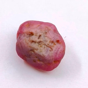 Puede incluir: Una gema de rubí en bruto sin cortar. El rubí tiene un tono rojo rosado intenso con áreas de color más claro y una superficie ligeramente texturizada. La piedra tiene una forma irregular, con una apariencia natural y orgánica.