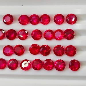 May include: A collection of twenty-four round, faceted, deep red gemstones arranged in four rows on a white surface. The gems have a brilliant shine, reflecting light. These gemstones are likely rubies, suitable for jewelry.