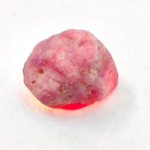 May include: A raw, unpolished gemstone with a vibrant pink and reddish hue. The stone features an irregular, rounded shape with a textured, matte surface and translucent edges that glow with a bright red light when illuminated from below.