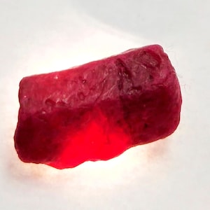 May include: A deep red ruby gemstone with a rough, natural shape. The ruby is illuminated from below, highlighting its rich colour.