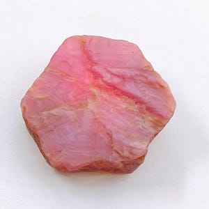 Puede incluir: Una piedra preciosa rosa en bruto de forma hexagonal con una superficie cristalina texturizada. La piedra muestra tonos variados de rosa claro y oscuro con estrías naturales visibles y áreas opacas y mate en su estructura irregular.