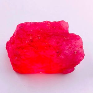 May include: A vibrant, rough-cut ruby gemstone. The ruby is a deep, saturated red color with a slightly uneven surface. The gemstone is displayed against a plain white background.