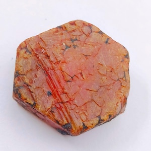 May include: A rough-cut, reddish-brown gemstone with a textured surface and dark veins. The stone has a hexagonal shape and displays a mix of red, brown, and black hues. The gemstone is likely a raw mineral specimen.