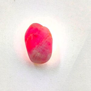 May include: A small, irregularly shaped gemstone with a vibrant, translucent hot pink hue. The stone features a smooth, polished surface with visible internal crystalline structures and a bright, glowing light effect emanating from within the center.