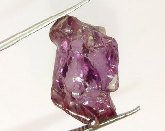 May include: A large, faceted, purple gemstone with a rough, natural finish. The gemstone is held in tweezers.