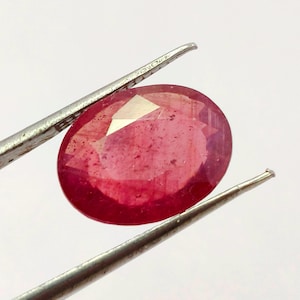 May include: A faceted, oval-shaped ruby gemstone, held by a pair of silver tweezers. The ruby displays a deep red hue with lighter internal patterns and facets that catch the light. The background is plain white.