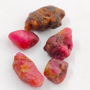 May include: Five rough, unpolished ruby gemstones in various shades of pink and red. The stones have a natural, raw appearance.