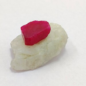 May include: A vibrant, fuchsia-colored gemstone rests atop a pale, off-white mineral. The gemstone has a rectangular shape with slightly rounded edges. The mineral base has a rough, uneven texture. The composition is set against a plain white background.