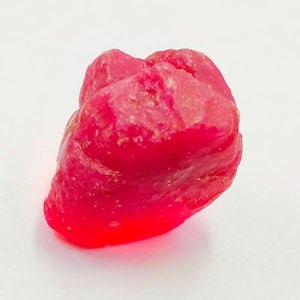 Puede incluir: Una gema de rubí en bruto sin cortar. El rubí es de un color rojo intenso y vibrante con una superficie rugosa y texturizada. La gema se encuentra sobre un fondo blanco liso.