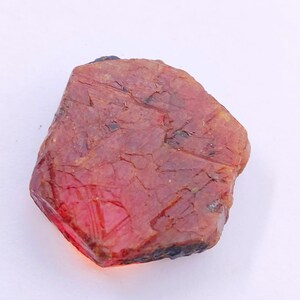 May include: A rough-cut, reddish-orange gemstone with a faceted, crystalline edge. The stone has a natural, textured surface with visible cracks and variations in color. The overall shape is irregular, showcasing its raw, unpolished state. This gemstone is ideal for jewelry making.