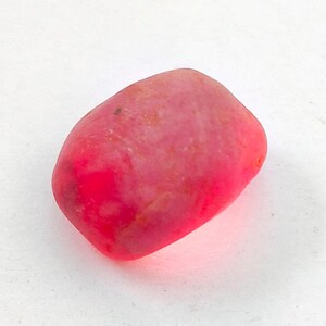 Puede incluir: Una piedra lisa y redondeada de color rojo, con aspecto pulido. La piedra tiene un efecto degradado, pasando de un rojo intenso a un tono rosa más claro. La forma es irregular, con bordes redondeados y una forma ligeramente alargada. Parece ser una gema natural.