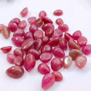 May include: A collection of polished, oval-shaped gemstones in shades of pink and red. The stones vary in size and have a smooth, glossy surface, reflecting light. The gemstones are scattered across a white surface.