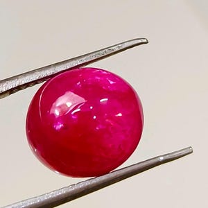 Könnte beinhalten: Ein leuchtend roter Cabochon-Edelstein, gehalten von einer Pinzette, mit einer glatten, polierten Oberfläche.