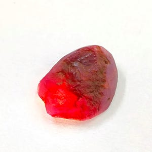 May include: A raw, unpolished gemstone with a vibrant red hue. The stone features an irregular, rounded shape with a textured, matte surface on the upper portion and a translucent, bright red crystalline section at the base.
