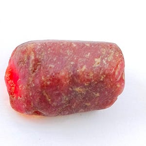 May include: A raw, unpolished gemstone with a cylindrical shape and a deep reddish-pink hue. The surface of the stone displays a natural, rugged texture with visible variations in colour intensity and opaque patches across its entire exterior.