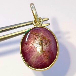 May include: A round cabochon ruby pendant set in a gold bezel with a small attached bail. The deep pinkish-red gemstone features visible internal inclusions and a smooth, polished surface, reflecting light across its rounded, dome-like top.