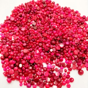 May include: A large collection of small, round, flat-backed beads in various shades of vibrant pink and magenta. The beads have a smooth, polished finish with subtle variations in color intensity and size, scattered across a plain white surface.