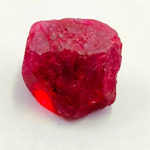 May include: A raw, uncut ruby gemstone with a deep red hue. The surface of the ruby has a rough, textured appearance, with areas of lighter and darker shades of red. The gemstone is set against a white background.