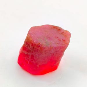 Puede incluir: Una gema de rubí en bruto, sin cortar, con un tono rojo vibrante. La gema tiene una superficie rugosa y texturizada y una forma natural e irregular. El color pasa de un rojo intenso en la base a un rojo rosado más claro en la parte superior. Esta gema es un mineral natural.