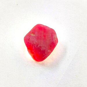 Puede incluir: Una pequeña piedra preciosa en bruto y sin tallar con un tono rosa rojizo vibrante y saturado. La piedra presenta una forma irregular con múltiples facetas, un aspecto semitranslúcido y una textura natural sin pulir que refleja la luz.