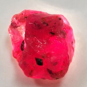 Op de afbeelding: Een grote, ruwe, roze robijn edelsteen met zwarte insluitsels. De robijn is een diepe, levendige roze kleur en heeft een glanzende afwerking.