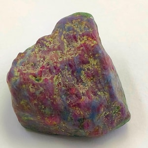 May include: A rough, heart-shaped ruby gemstone with a mix of red, pink, green, and blue hues. The stone has a natural, unpolished finish.