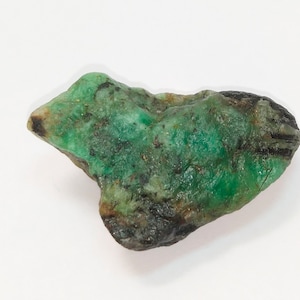 May include: A rough, green emerald gemstone with black inclusions. The emerald is a natural, unpolished stone.