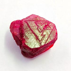 Puede incluir: Una gema de rubí en bruto y sin cortar, de color rojo intenso. La superficie presenta un patrón único de inclusiones doradas, creando un contraste llamativo. La gema tiene forma irregular, mostrando su estado natural y sin pulir.