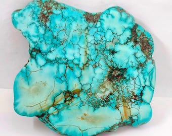 Pode incluir: Uma vibrante pedra preciosa turquesa com uma forma irregular única. A pedra apresenta uma rede de veias e fissuras castanho-escuras, criando um contraste marcante. As áreas polidas da pedra refletem a luz, realçando a sua beleza natural.