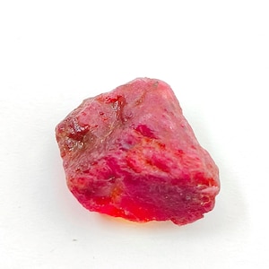 May include: A natural, rough-cut ruby gemstone. The ruby is a deep red colour with a slightly translucent appearance. The surface has a textured, uneven appearance, with visible crystalline structures. The gemstone is set against a white background.