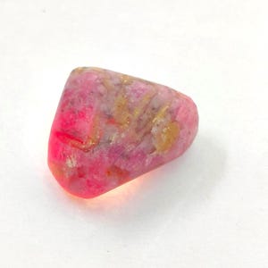 Op de afbeelding: Een gepolijste, onregelmatig gevormde edelsteen met een levendige mix van felroze, magenta en zachte goudgele tinten. De steen heeft een glad, afgerond oppervlak met zichtbare interne kristalstructuren en een gevarieerde kleurverzadiging.