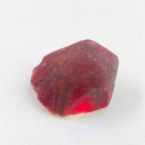 May include: A raw, uncut gemstone with a deep red colour and a textured, irregular surface. The stone displays a translucent quality with a bright, concentrated red glow visible near the bottom edge, showcasing natural facets and mineral inclusions.