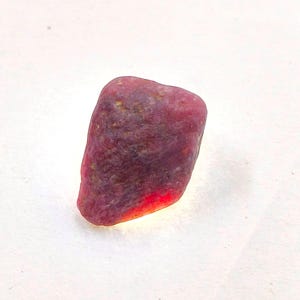 May include: A raw, unpolished ruby gemstone with a deep red and magenta hue. The stone features an irregular, naturally textured surface with visible dark inclusions and a translucent, glowing base that catches light, showing a vibrant crimson tone.