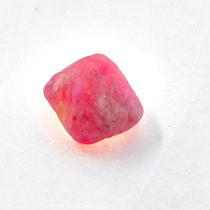 May include: A small, rough, and irregularly shaped stone with a vibrant hot pink colour. The surface of the mineral displays a textured, matte appearance with subtle variations in tone, featuring patches of lighter white and deeper pink hues.
