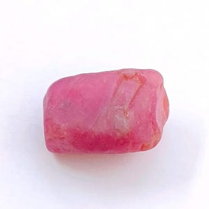 May include: A rough-cut, pink gemstone with a smooth, polished surface. The stone is rectangular and has a vibrant, saturated colour. The gemstone is set against a white background.