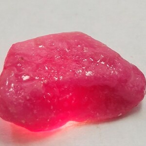 Puede incluir: Una gran gema rosa rugosa con una superficie lisa y pulida. La gema está iluminada desde abajo, lo que resalta su calidad translúcida.