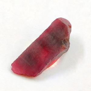 May include: A raw, elongated ruby gemstone with a deep red color and translucent qualities. The stone features a natural, uneven surface texture with visible internal inclusions and a slightly tapered shape, showcasing a vibrant crimson hue.