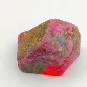 Op de afbeelding: Een ruwe, ongeslepen robijn edelsteen met een levendige roze tint. De steen heeft een textuur met bruine, grijze en rode gebieden. De robijn staat tegen een witte achtergrond, wat de natuurlijke schoonheid benadrukt.