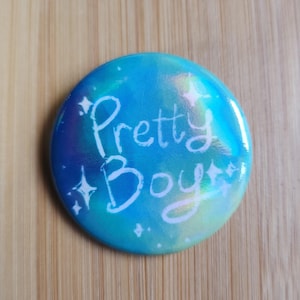 Puede incluir: Un botón redondo con un fondo azul y turquesa y las palabras "Pretty Boy" en escritura blanca. El botón tiene un acabado brillante y detalles de estrellas.