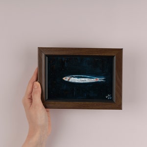 May include: A framed painting of a silver fish on a dark blue background. The painting is in a dark brown frame and has the signature 'OK 23' in the bottom right corner. Sardine art original painting Sardines still life oil painting miniature Anchovy fish painting French country kitchen art 4x6