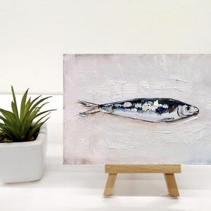 May include: An oil painting of a fish on a white background, displayed on a small wooden easel. The fish is painted in shades of black, white, and silver, with visible brushstrokes. A small potted plant is to the left of the painting.
