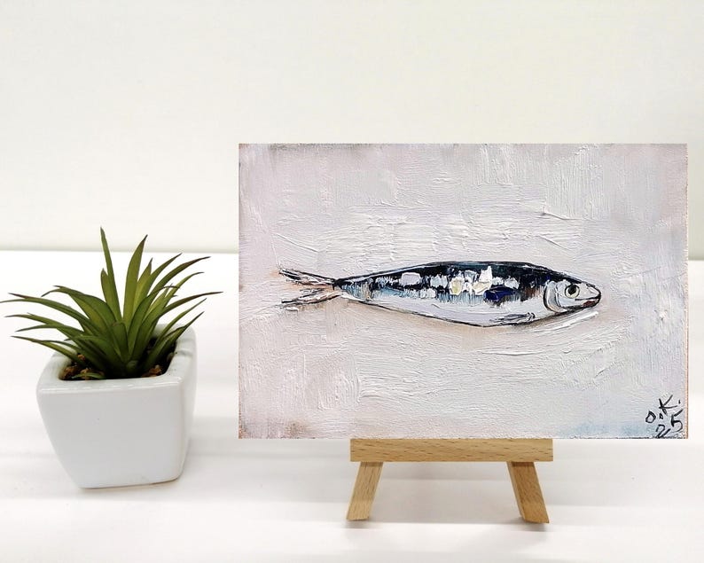 May include: An oil painting of a fish on a white background, displayed on a small wooden easel. The fish is painted in shades of black, white, and silver, with visible brushstrokes. A small potted plant is to the left of the painting.