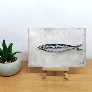 May include: A small painting of a single silver fish on a white background. The painting is on a small easel and has the signature "OK 22" in the bottom right corner.