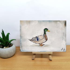 May include: A small painting on a wooden easel of a duck with a green head and a yellow beak. The duck is standing on a white background. Mallard duck painting original miniature art Vintage duck oil painting farmhouse kitchen wall art country decor duck hunting gift 4x6