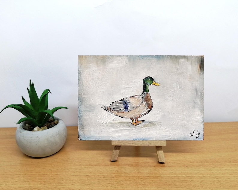 May include: A small painting on a wooden easel of a duck with a green head and a yellow beak. The duck is standing on a white background. Mallard duck painting original miniature art Vintage duck oil painting farmhouse kitchen wall art country decor duck hunting gift 4x6