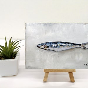 May include: A small painting on a wooden easel depicts a single, silver-colored fish with blue and black markings on a white background. The painting is signed "OK 22" in the bottom right corner.