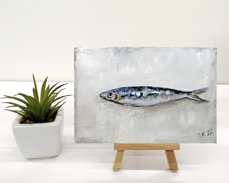 May include: A small painting on a wooden easel depicts a single, silver-colored fish with blue and black markings on a white background. The painting is signed "OK 22" in the bottom right corner.