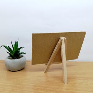 May include: A small, unfinished wooden easel with a rectangular, cardboard-like surface. The easel is propped up on a wooden table, with a small potted plant to the left. The background is a plain white wall.
