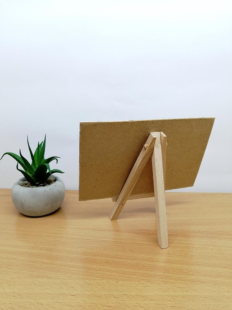 May include: A small, unfinished wooden easel with a rectangular, cardboard-like surface. The easel is propped up on a wooden table, with a small potted plant to the left. The background is a plain white wall.