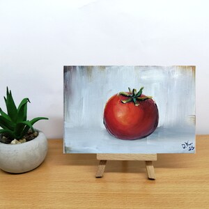 May include: A small painting of a red tomato on a white background. The painting is on a small easel.